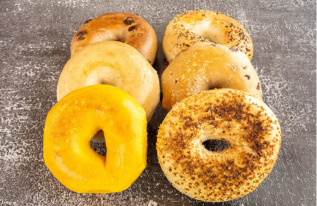 Six assorted bagels arranged in two rows on a textured gray surface, featuring different toppings and flavors including cheese, seeds, and plain.