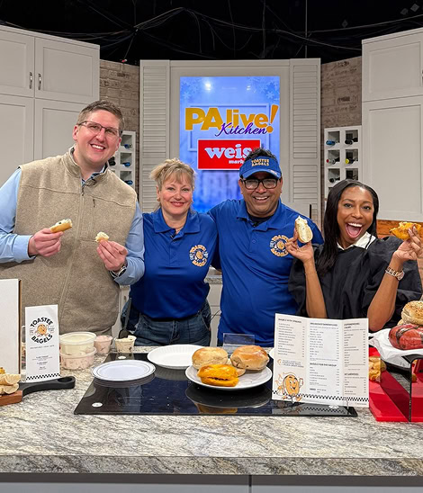 Four people stand behind a kitchen counter holding bagels and smiling during a TV cooking segment with a “PA live! Kitchen” sign in the background.