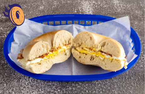 A sliced bagel sandwich with egg and bacon is placed in a blue plastic basket lined with wax paper, on a textured gray surface.