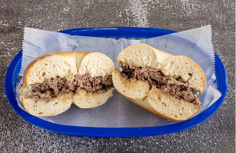A sliced bagel filled with shredded beef and melted cheese sits on parchment paper in a blue plastic tray on a textured surface.