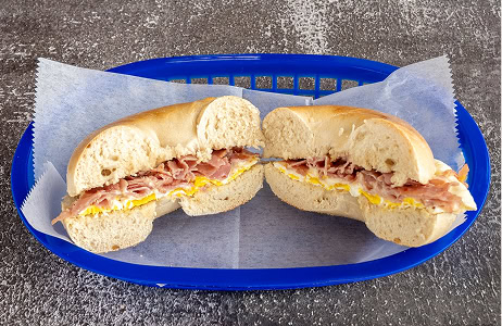 A bagel sandwich with scrambled eggs and sliced ham, cut in half and served on parchment paper in a blue plastic basket.