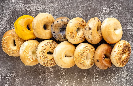 Twelve assorted bagels, including plain, sesame, poppy seed, everything, blueberry, and cheese, arranged in two neat rows on a textured gray surface.