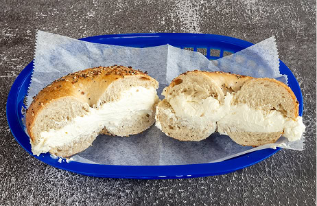 A sliced everything bagel with cream cheese is placed in a blue plastic basket lined with parchment paper on a gray textured surface.