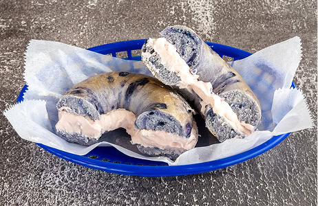 A sliced purple bagel with cream cheese filling is placed in a blue basket lined with parchment paper on a textured surface.
