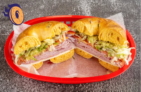 A sandwich made with an onion bagel, filled with sliced deli meat, lettuce, and tomato, cut in half and served in a red plastic basket lined with parchment paper.