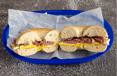 A bagel sandwich cut in half, filled with scrambled eggs and sliced meat, served on wax paper in a blue plastic basket.