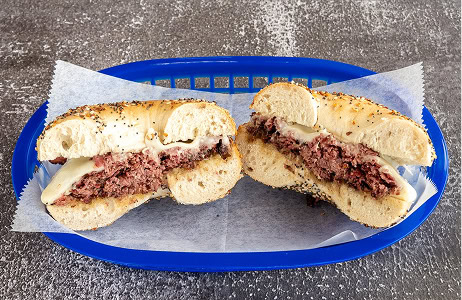 A sliced everything bagel with a beef patty and melted cheese is served in a blue plastic basket lined with parchment paper on a gray textured surface.