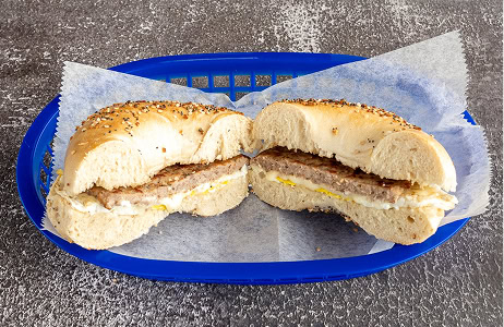 A breakfast sandwich with sausage, egg, and cheese on a seeded bagel, cut in half and served on wax paper in a blue basket.