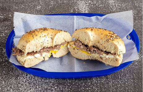 Everything bagel sandwich with meat, egg, and mustard, cut in half and served on parchment paper in a blue basket on a textured surface.