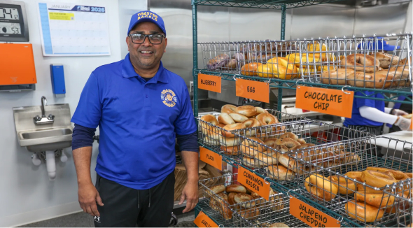 A man wearing a blue branded polo shirt and cap smiles next to wire shelves filled with labeled bins of bagels.